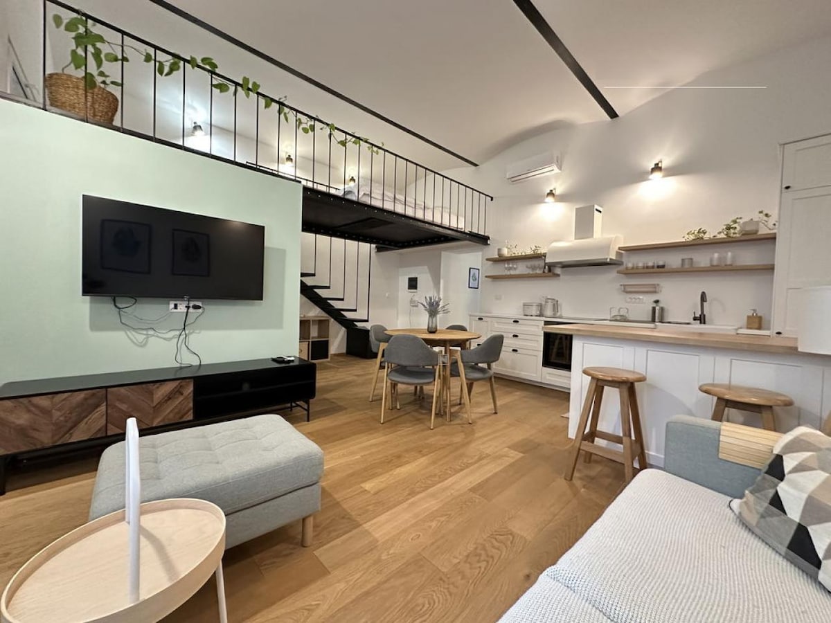 The spacious living area features natural wood flooring and a mix of modern furnishings. A large television is mounted on the wall, opposite a light-colored sofa and a small coffee table. The open kitchen, equipped with various appliances, is enhanced by a mezzanine sleeping area visible above.