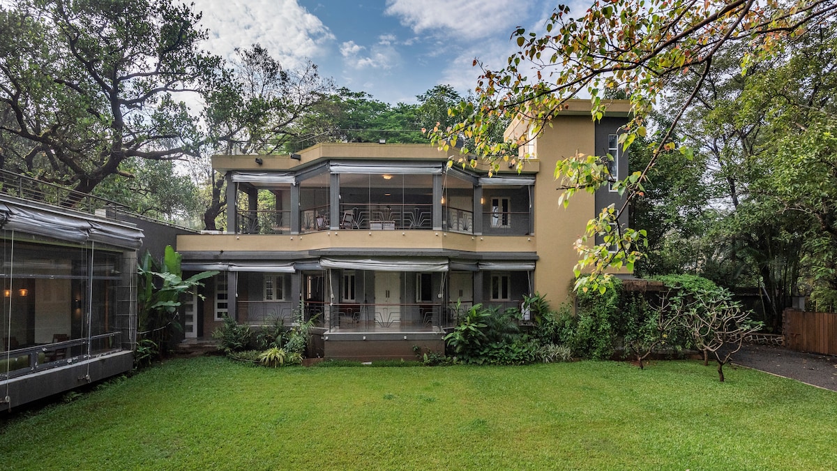 The villa is set within a lush green lawn, surrounded by greenery and trees. A two-story building showcases a spacious verandah on the ground floor and multiple balconies above. Large windows invite natural light, while the tranquil outdoor space offers a serene environment.