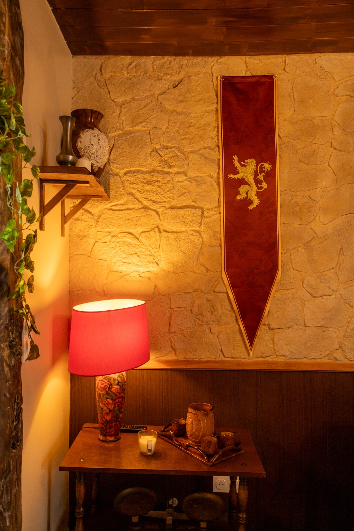 A warm corner of the room is highlighted by a red lamp, softly illuminating a wooden table. Textured stone walls are featured, adorned with a decorative banner displaying a lion motif. A few rustic items and a candle are placed on the table, adding character to the space.