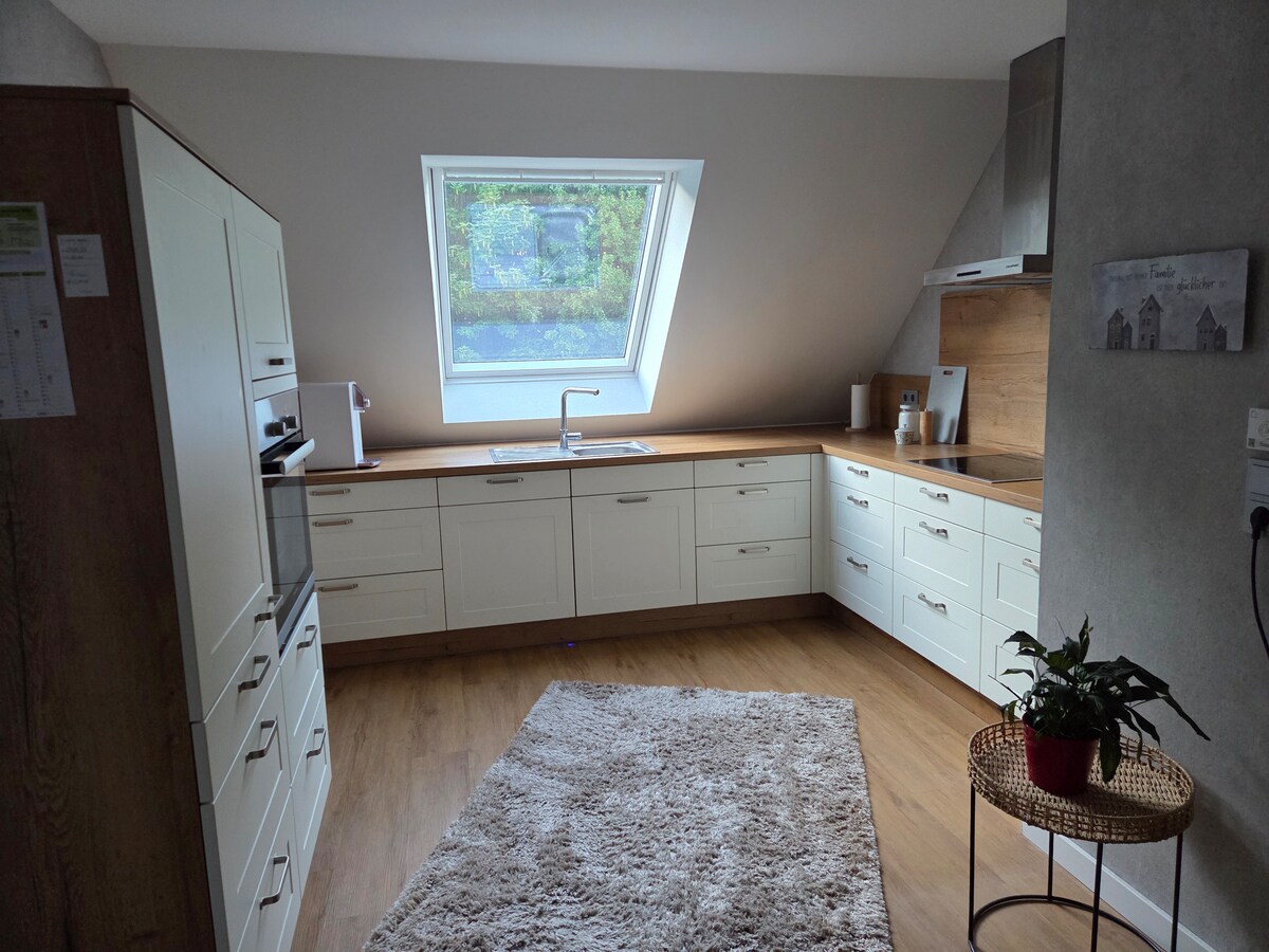 The kitchen features modern white cabinetry with ample storage, complemented by a warm wooden countertop. A large slanted window allows natural light to enter, while a small potted plant adds a touch of greenery. A rug enhances the comfort of the space.