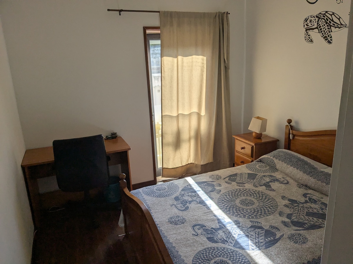 A private bedroom features a spacious bed adorned with a patterned blanket. Natural light filters through a curtain covering the window. A wooden desk and chair are positioned nearby, along with a small nightstand. The walls are painted in a light hue, contributing to a calm ambiance.