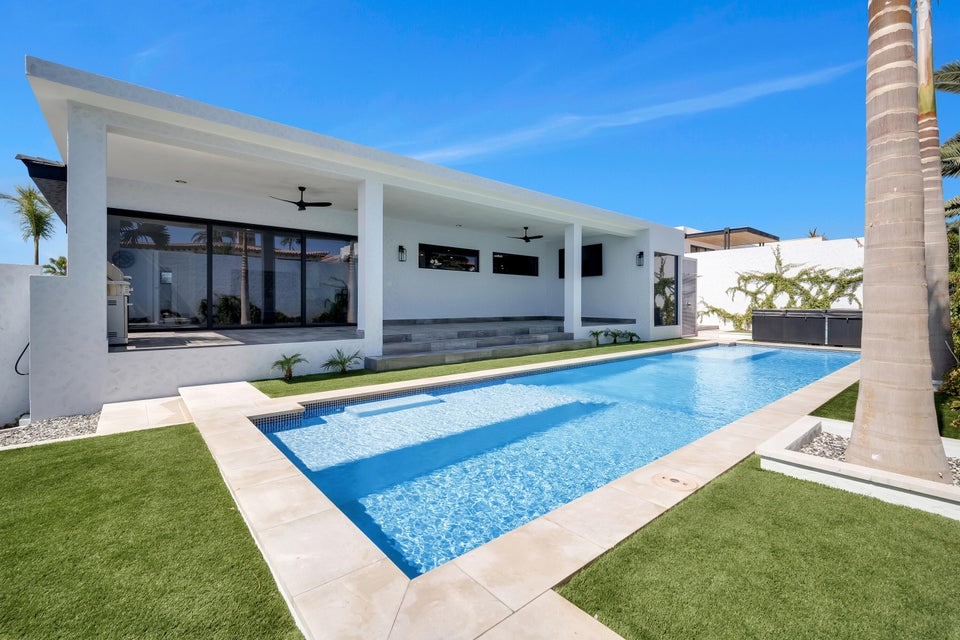 A spacious outdoor area showcases a long swimming pool framed by elegant tilework and lush green grass. The modern architecture features expansive glass doors and covered patio spaces, offering a seamless connection to the surrounding landscape. Palm trees enhance the tropical ambiance.
