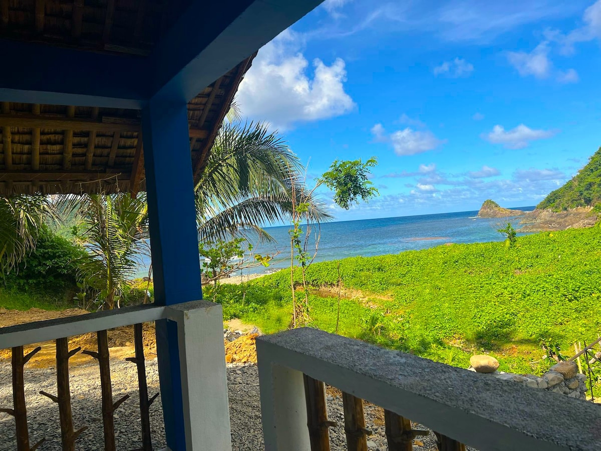 A serene coastal view is captured from a shaded porch, framed by a thatched roof. Lush greenery extends towards the water, showcasing an inviting shoreline under a clear blue sky. Gentle waves can be seen lapping against rocks in the distance.