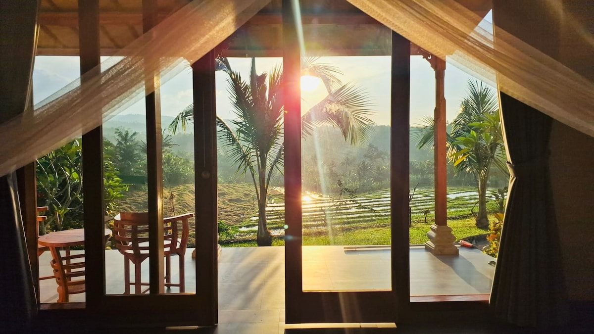 A serene view is captured through large glass doors, revealing a sunlit landscape. Lush palm trees and rice fields are visible, while a wooden table and chairs are positioned on a spacious terrace, inviting relaxation in a tranquil setting.