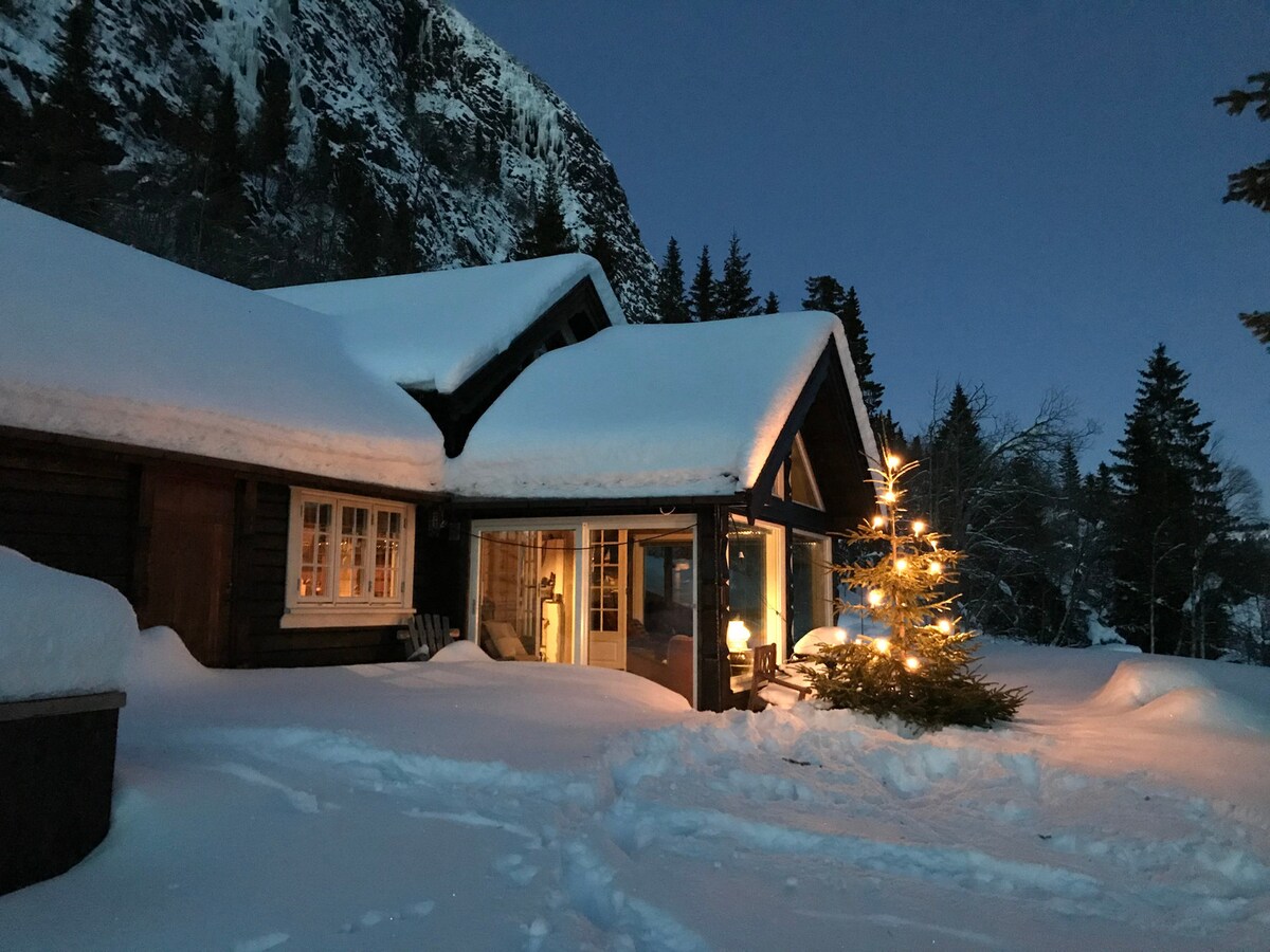 A cozy cabin is enveloped in a blanket of snow, with a beautifully adorned Christmas tree positioned by the entrance. Warm light glows from the windows, casting a welcoming ambiance against the backdrop of frosty trees and towering mountains.