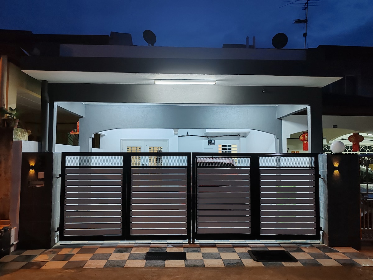 The exterior showcases a double-storey house viewed from the entrance, featuring a modern designed gate with horizontal slats. Soft lighting highlights the pathway leading to the front door, while the facade is complemented by a covered porch, creating an inviting entryway.