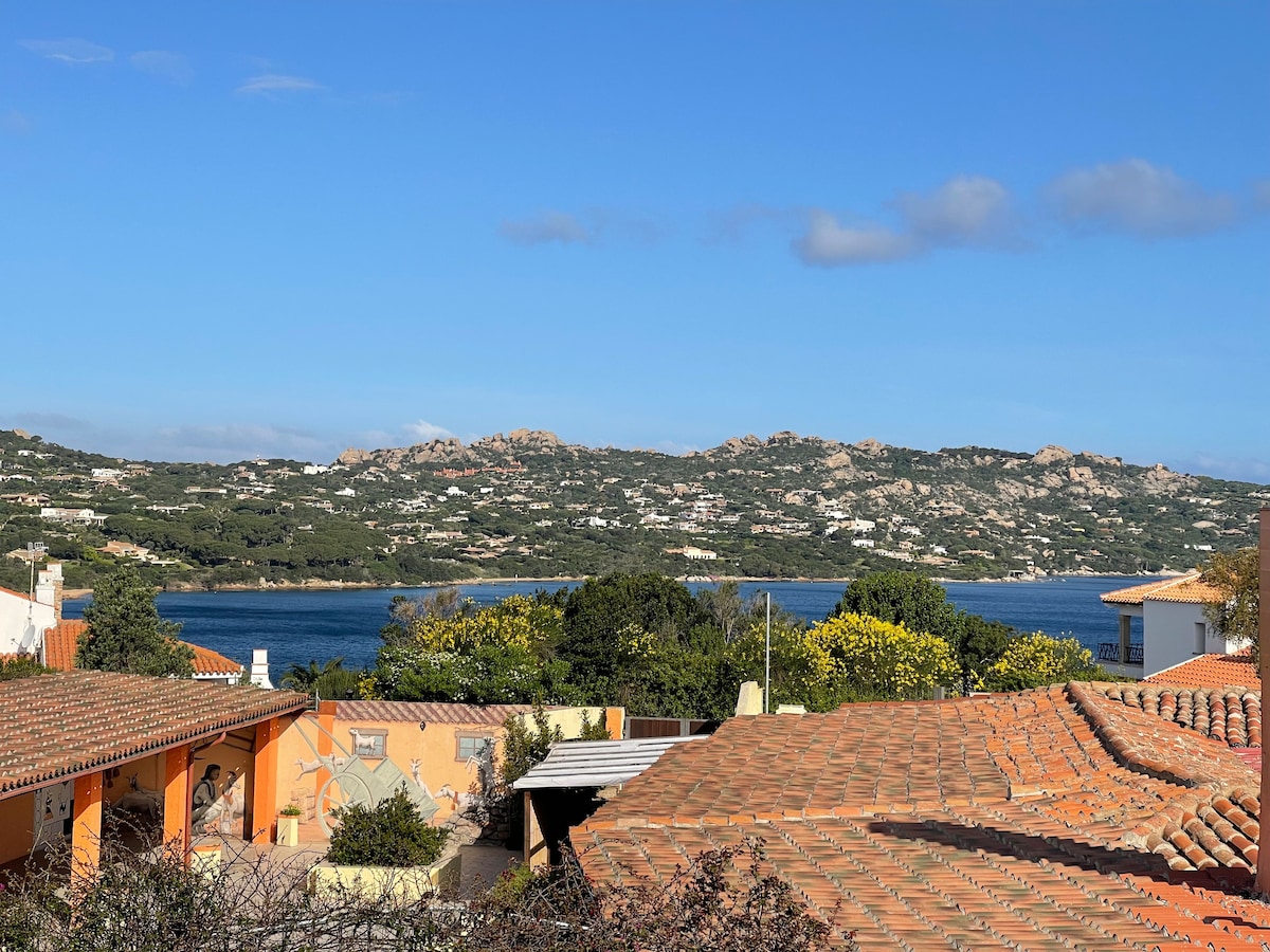 A panoramic view of the coastline is captured, highlighting blue waters and green hills in the distance. The foreground includes terracotta rooftops, providing a warm contrast to the scenery. A clear sky is visible, suggesting a bright and pleasant atmosphere.