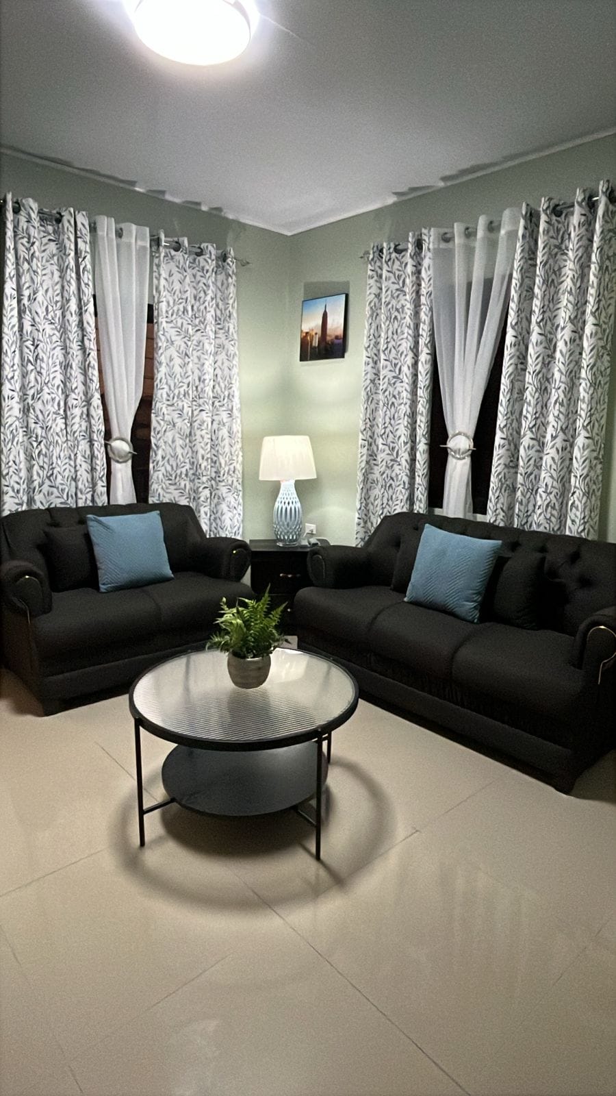 A seating area is depicted with two dark sofas adorned with light blue cushions, arranged around a circular glass table. A small potted plant sits on the table. Sheer curtains frame the windows, allowing natural light to filter through. A lamp provides additional illumination.