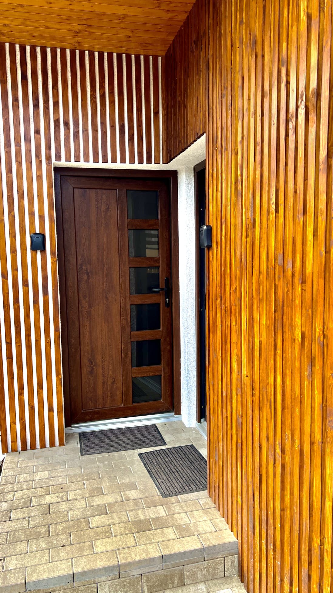 The entrance features a wooden door with glass panels, set against a backdrop of vertically striped wood. Light-colored tiles lead to the door, complemented by textured doormats on either side. The overall design conveys a modern and natural appeal.
