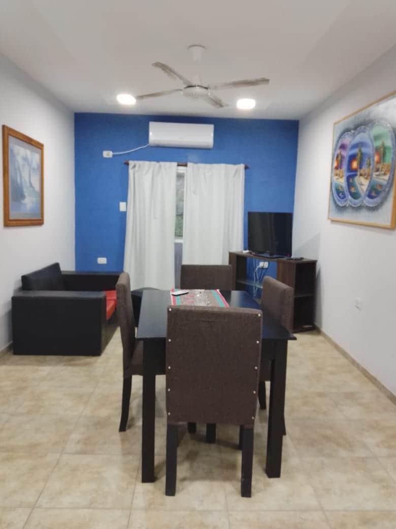 A living and dining area features a dark wood dining table surrounded by four chairs. A black sofa is positioned alongside a vibrant red cushion. Light-colored walls are complemented by blue accents, with a ceiling fan and a television mounted on a wooden stand.