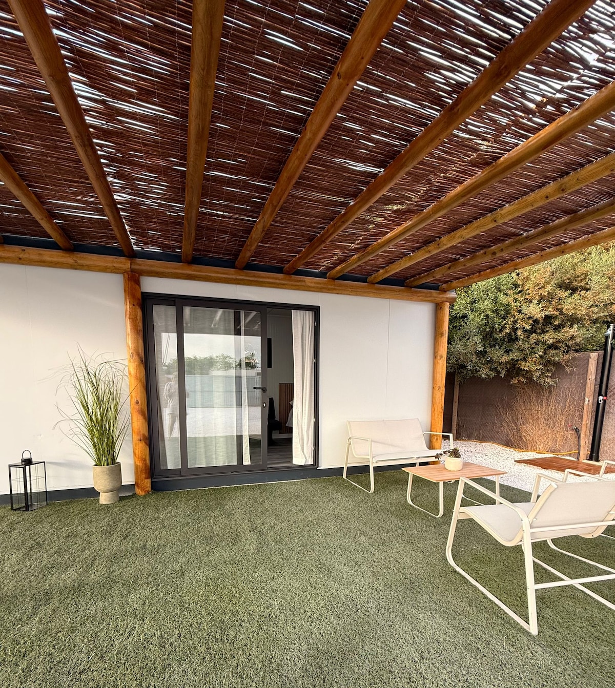 An outdoor seating area is presented under a woven roof, providing shade. Two light-colored chairs are arranged around a small table, accompanied by a potted plant. The green grass-like flooring enhances the relaxing setting, with a glimpse of foliage visible in the background.