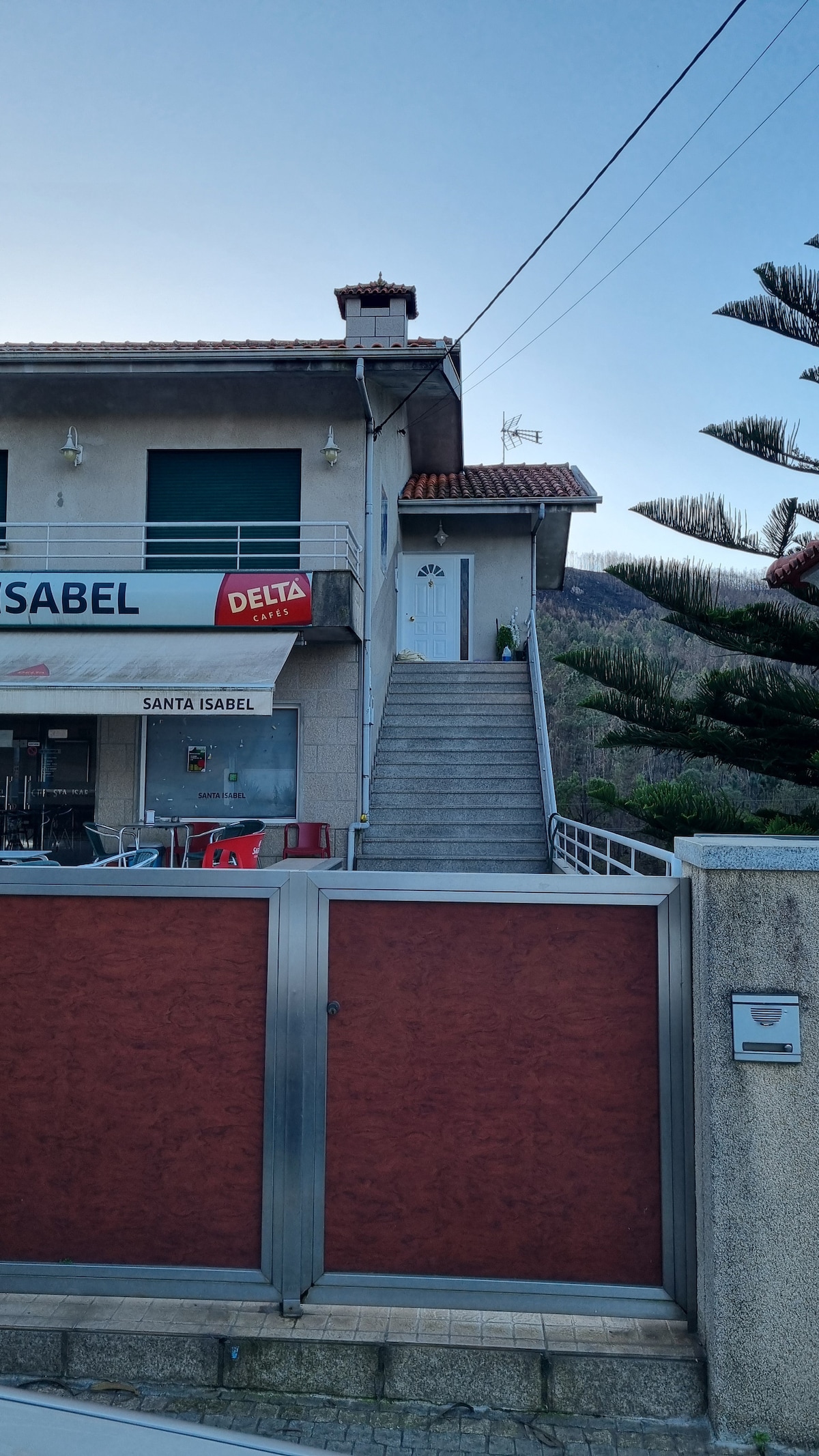 A two-story building is presented with a set of stairs leading to an entrance marked by a white door. The exterior is complemented by a sign that reads 'Santa Isabel'. A red and gray gated fence frames the property, with a nearby shrubbery adding to the landscape.