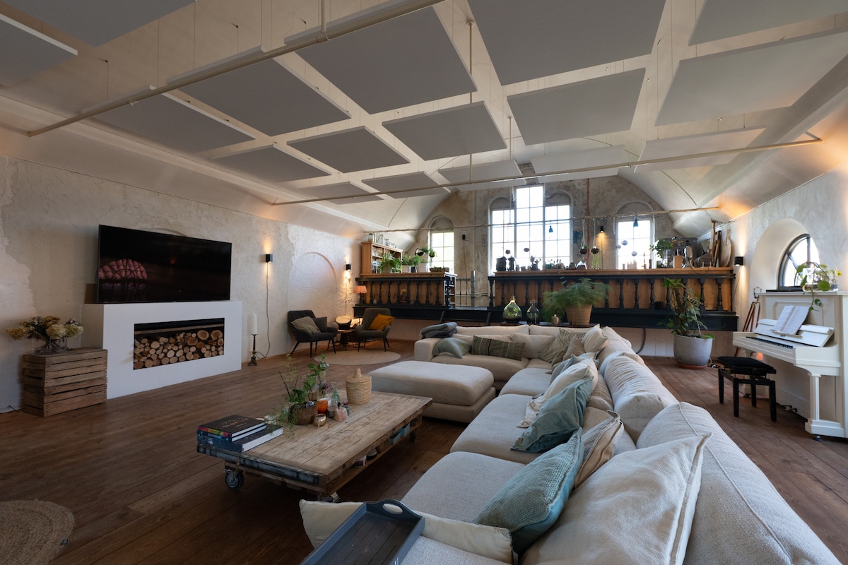 A spacious living area features high vaulted ceilings, large windows that provide ample natural light, and a cozy sectional sofa arranged around a wooden coffee table. A flat-screen TV is mounted on the wall above a white fireplace, with plants and decorative elements adding a touch of greenery.