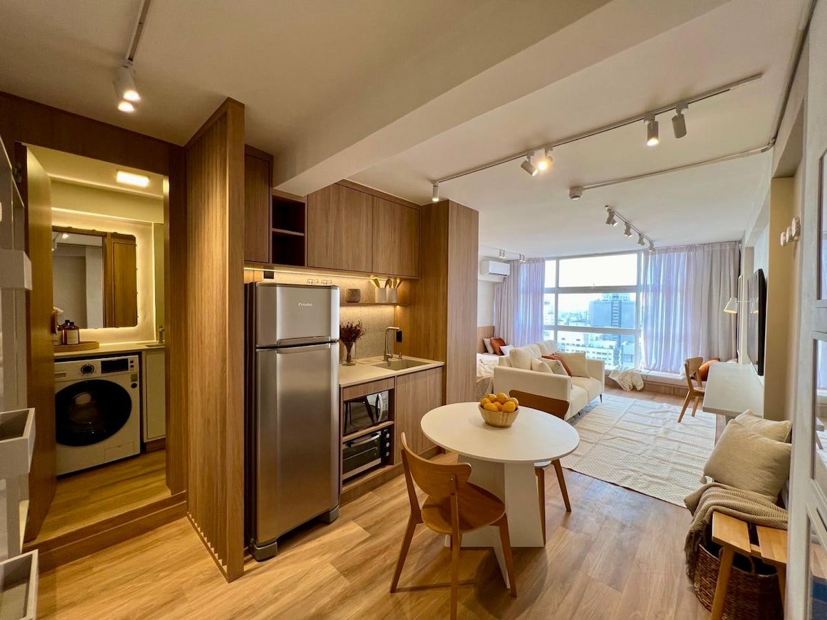 An open-concept studio is displayed, featuring a kitchen with modern appliances, including a refrigerator and stovetop. A round dining table with wooden chairs is positioned beside a living area, which is adorned with a light-colored rug and a large window offering city views.