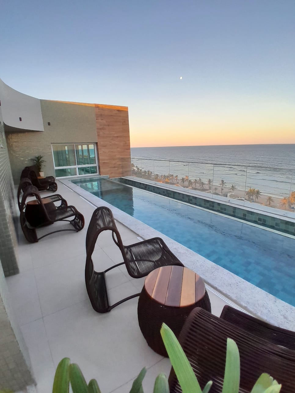 An infinity pool overlooks the ocean, creating a seamless view of the horizon. Lounge chairs are arranged nearby, providing space for relaxation. The warm colors of the sunset reflect off the water, enhancing the tranquil atmosphere of the rooftop area.