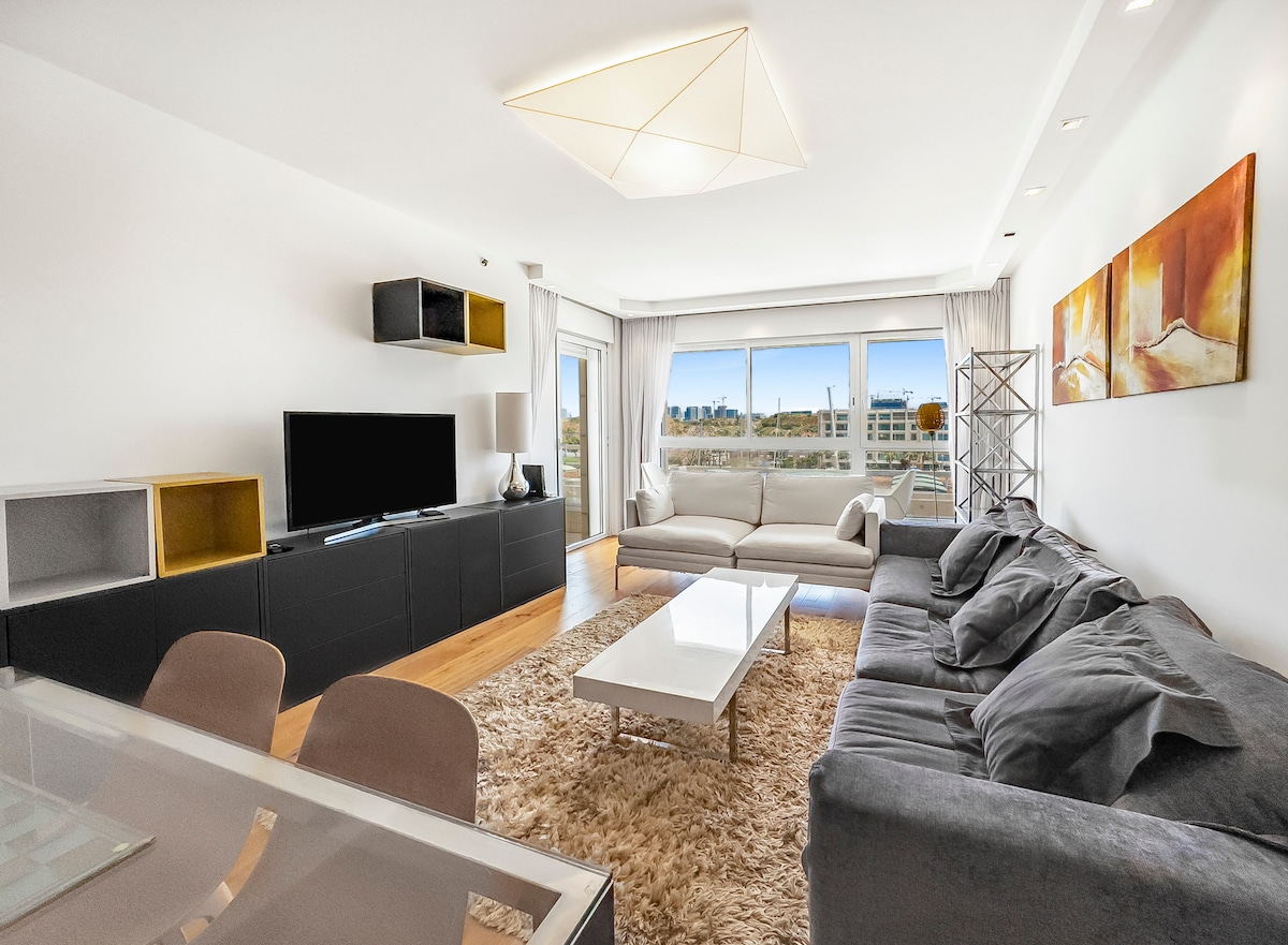 A spacious living area features a large, comfortable seating arrangement with soft grey upholstery. A modern coffee table sits at the center on a light rug, while a sleek TV unit stands opposite. Large windows offer natural light and views of the surrounding area.