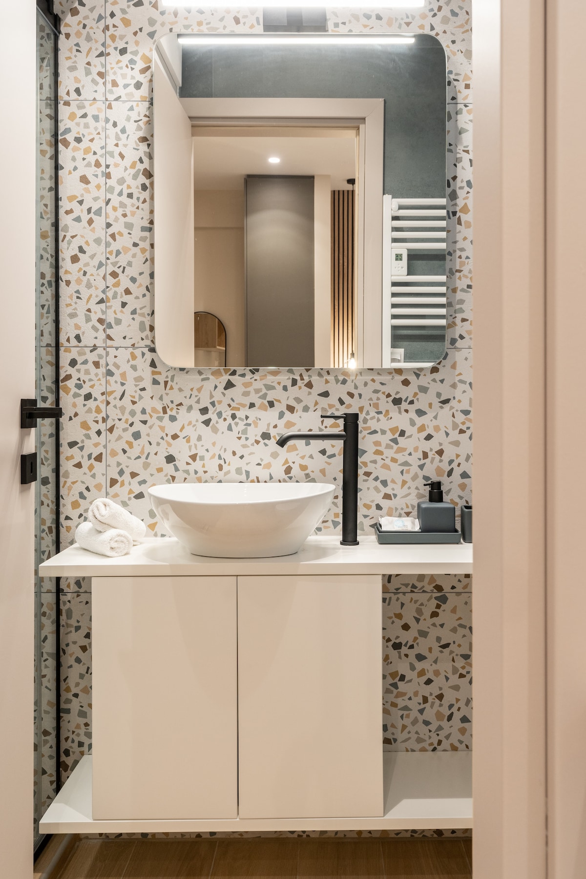 A modern bathroom is highlighted by a stylish sink and a large mirror. The walls feature a decorative patterned tile design. Fresh towels are neatly arranged beside the sink, which is complemented by a sleek faucet and countertop accessories.