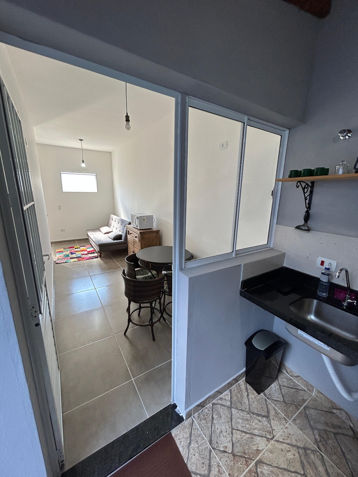 A view from the kitchen area shows a small dining table with two chairs, connected to a living space. Natural light enters through a window, illuminating a cozy seating arrangement in the background. A countertop includes basic kitchen amenities beside a trash bin.