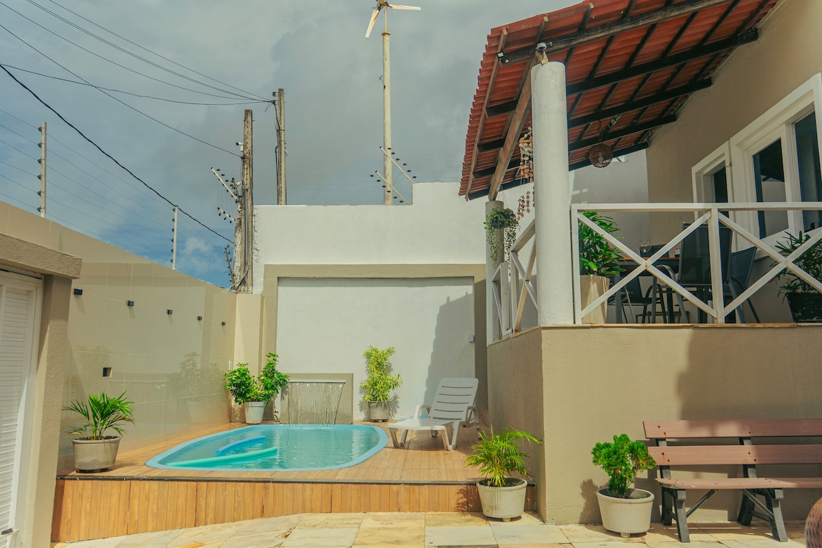 An outdoor area features a small pool surrounded by wooden decking. Potted plants add greenery, and a shaded patio with a slanted roof provides seating options. A comfortable lounge chair is positioned nearby, creating a relaxing atmosphere.