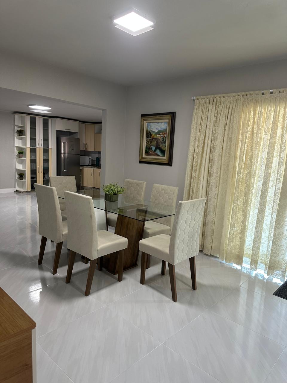 A dining area is displayed, featuring a glass table surrounded by six upholstered chairs. Natural light filters through sheer curtains, illuminating the space. A small potted plant rests at the center of the table, adding a touch of greenery to the neutral decor.