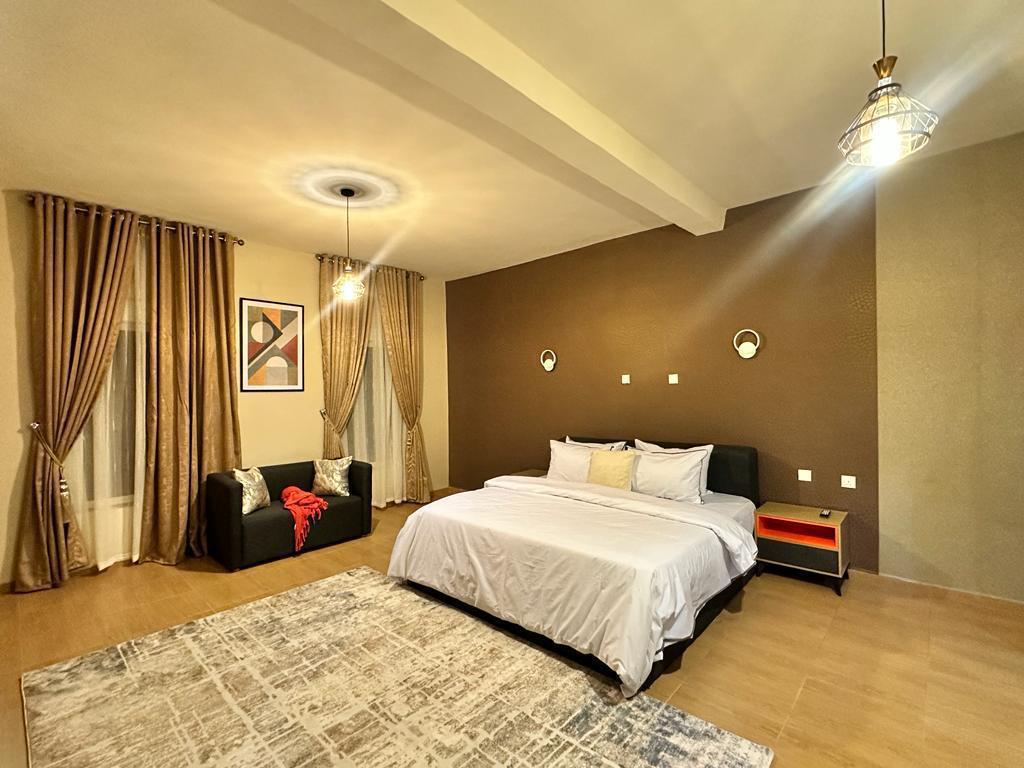 An inviting bedroom is depicted, featuring a spacious bed adorned with neutral linens. A large rug lies on the floor, complemented by a sitting area with a dark sofa. Soft lighting is provided by two pendant fixtures, and elegant curtains frame large windows.
