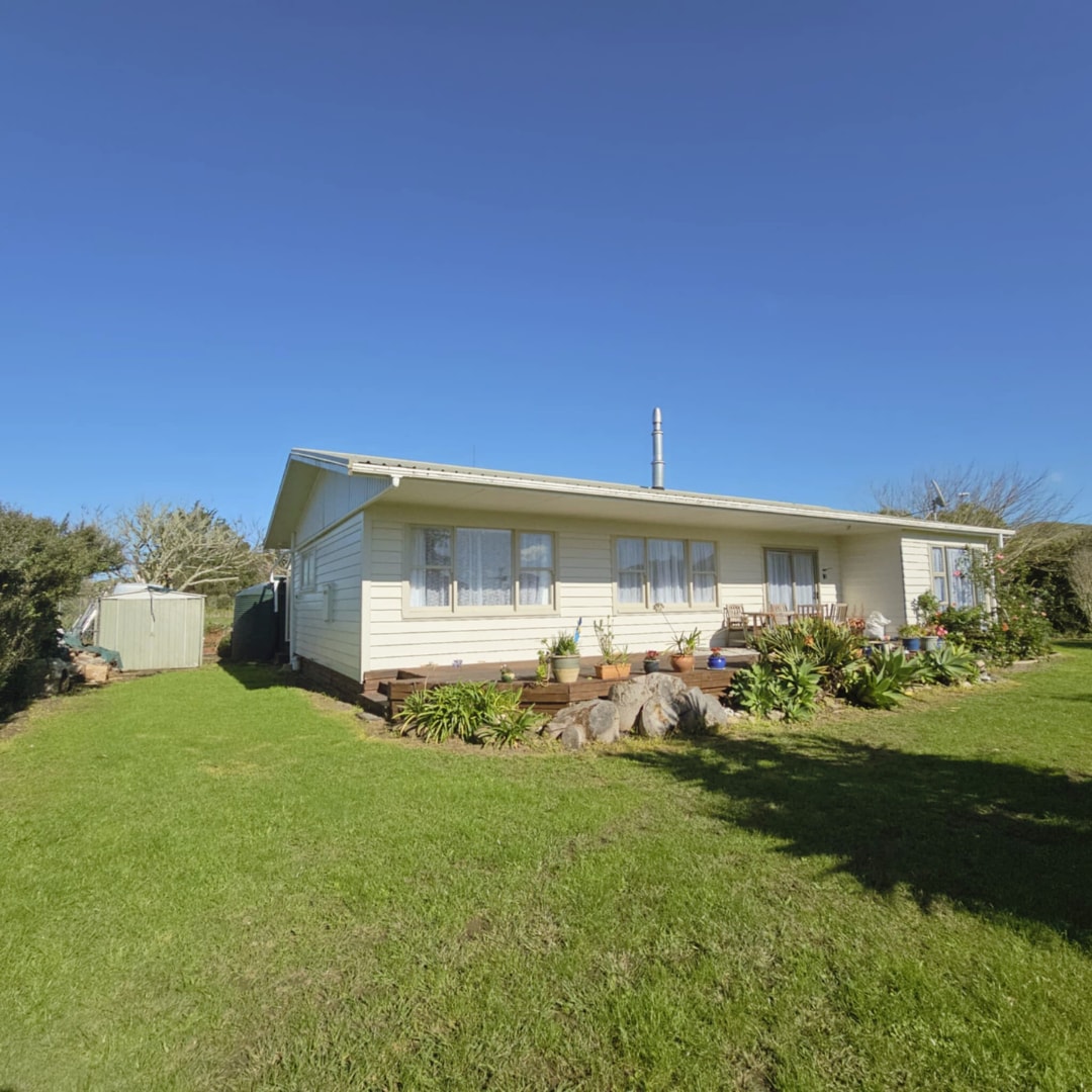 A single-story home is set against a clear blue sky, surrounded by lush green grass and well-kept gardens. The inviting exterior features multiple windows and a wooden porch area, with a small shed visible to the left.