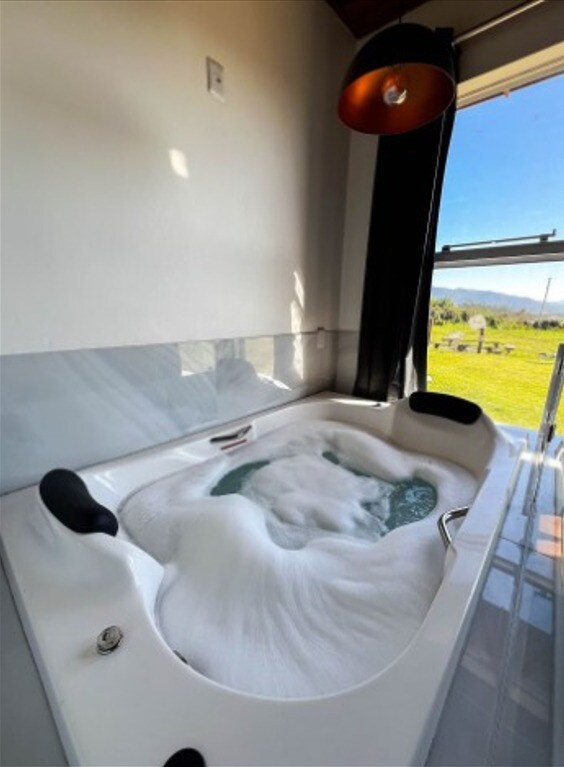 A modern jacuzzi is positioned beside a large window, with sunlight illuminating the bubbles. A scenic view of the grassy landscape is visible outside, while a stylish pendant light adds brightness to the space.