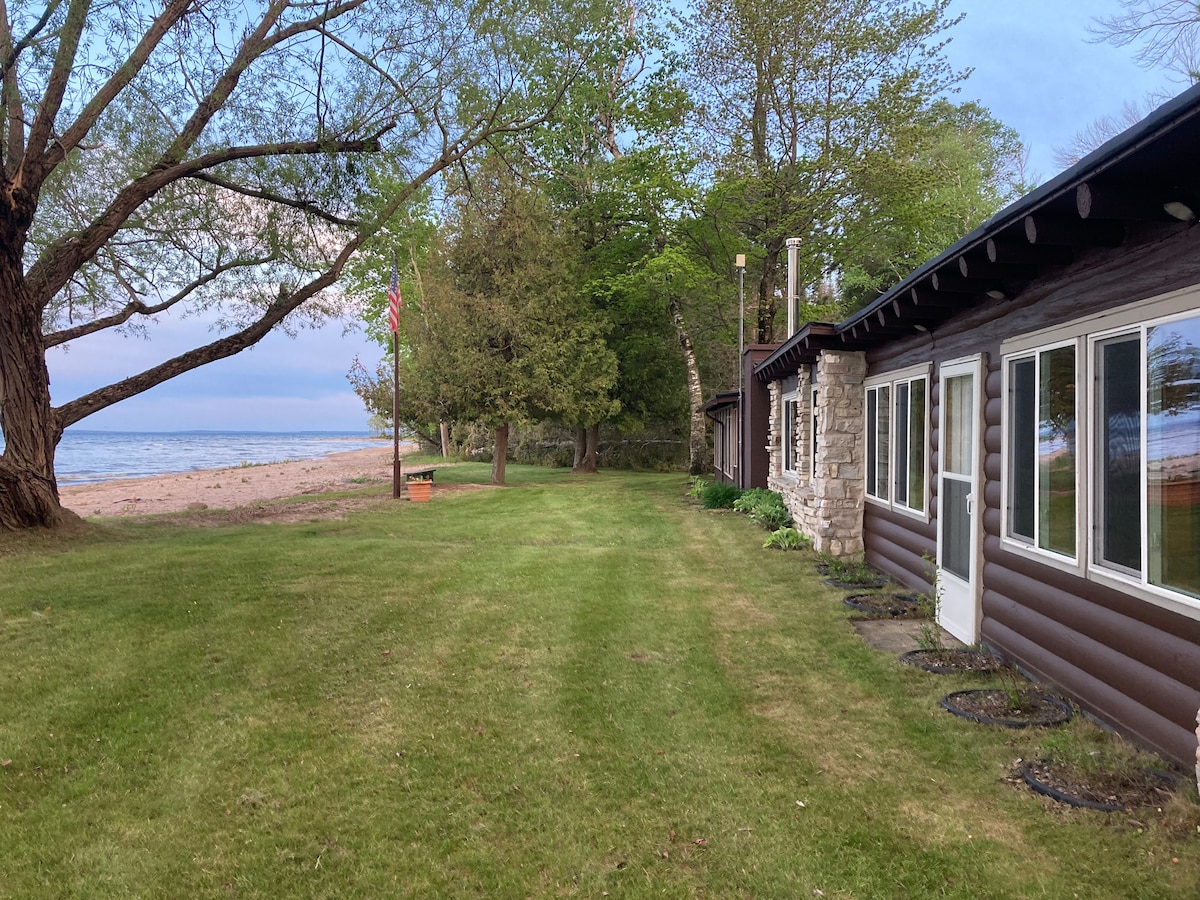 A well-maintained lawn extends alongside a charming cabin, offering direct access to a sandy beach. Large windows adorn the cabin's exterior, providing a view of Lake Michigan. Lush green trees frame the scene, contributing to a serene outdoor atmosphere.