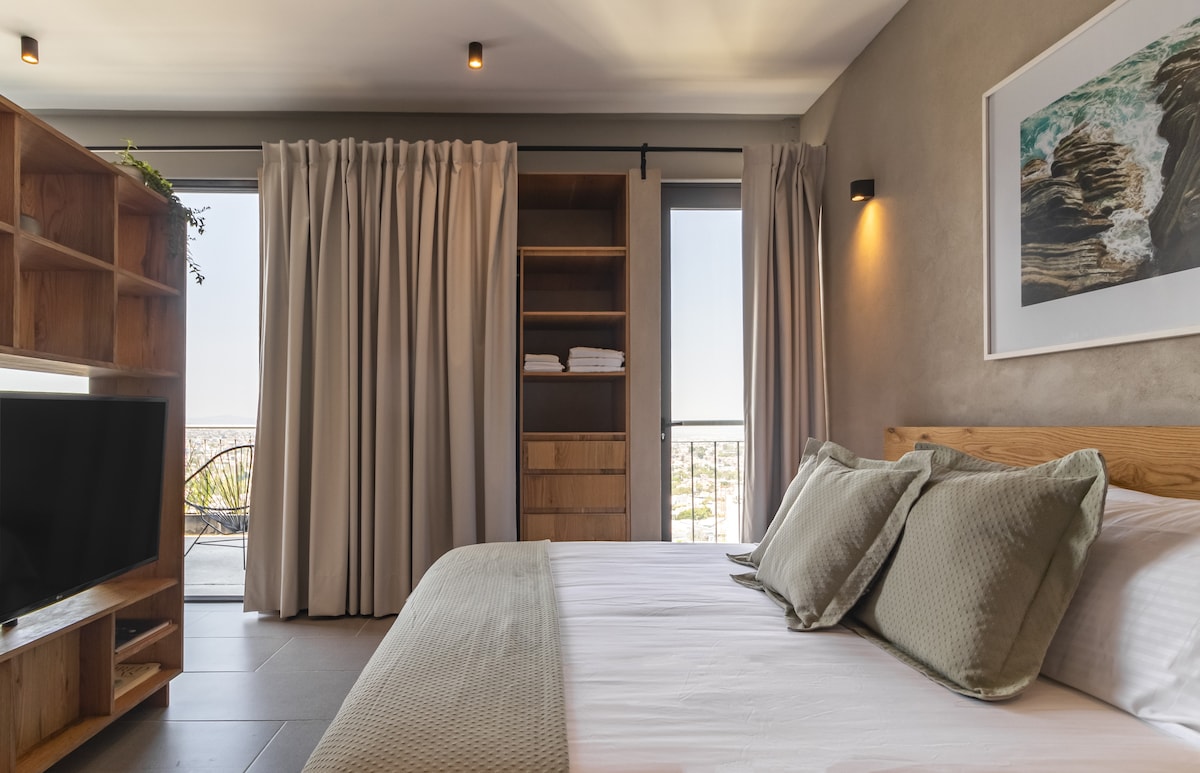 A modern bedroom features a spacious bed with muted green accents. Soft drapes frame the large windows, revealing a skyline view. A wooden shelving unit, with towels neatly stored, complements the décor. A television is positioned on a low stand for convenient viewing.