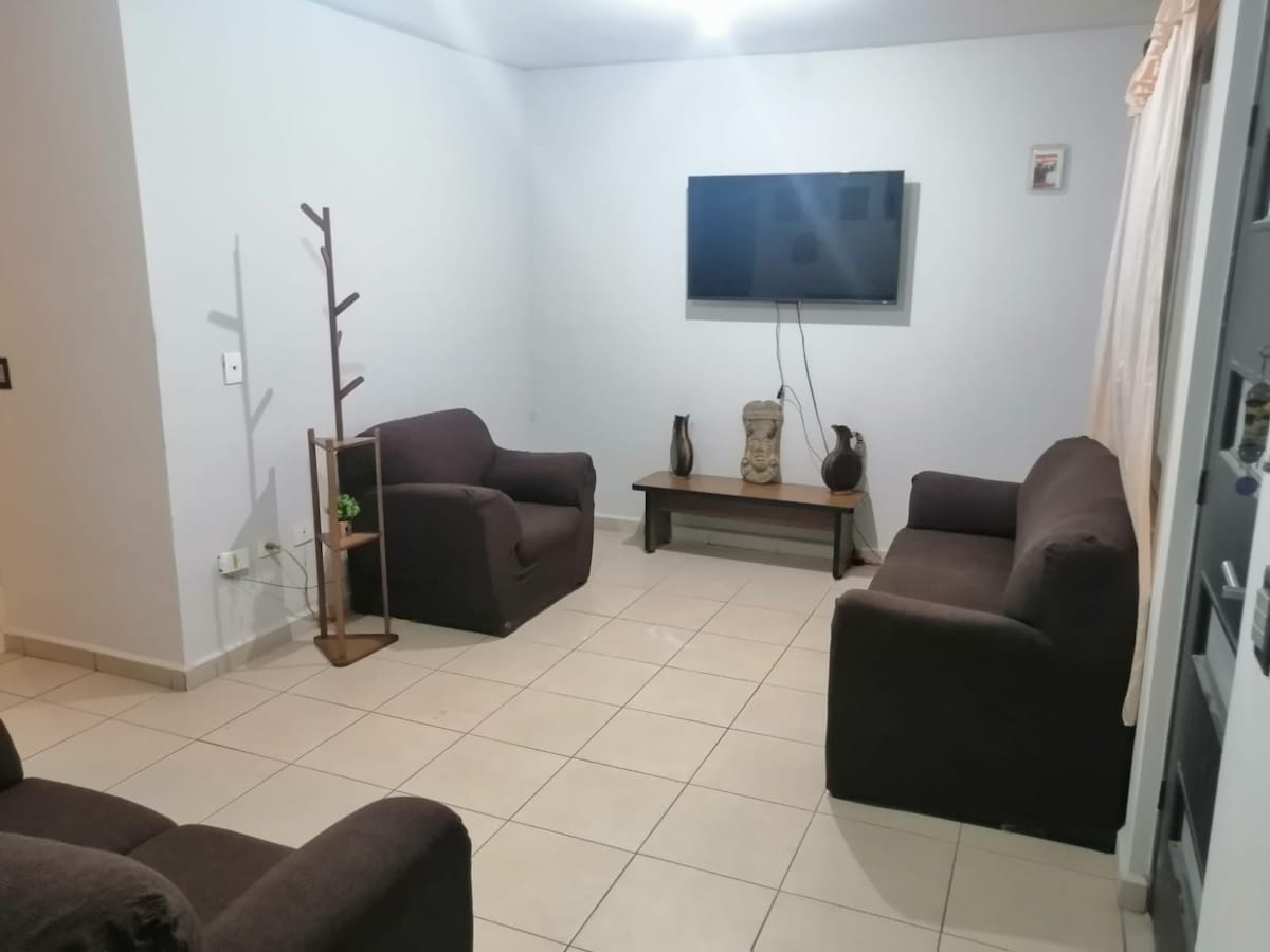 A living area features two dark brown sofas positioned opposite each other. A wooden coffee table sits at the center, accompanied by decorative vases. A flat-screen television is mounted on the wall, providing a focal point in the space. Soft lighting enhances the room's inviting setup.