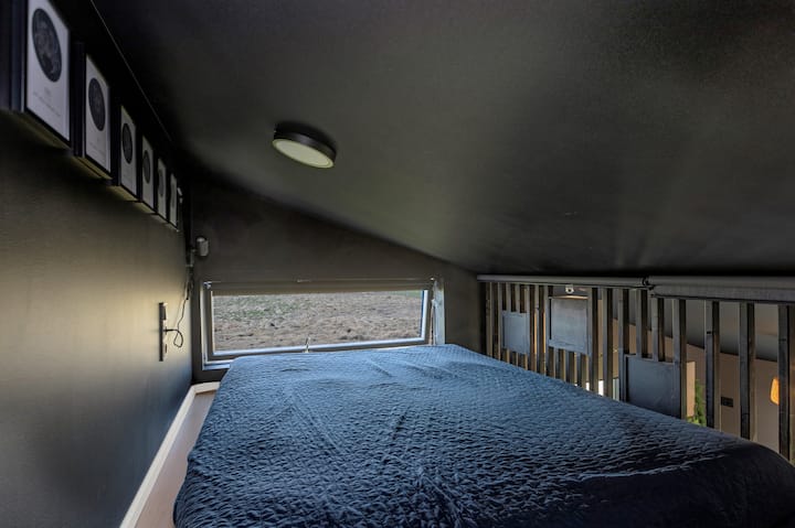 Este acogedor dormitorio tipo loft en la casa de invitados tiene el techo bajo, las paredes oscuras, una gran ventana con vistas panorámicas y una cama para dos. Las obras de arte enmarcadas y los detalles de las barandillas añaden un toque moderno e íntimo, perfecto para una noche de descanso tranquila.