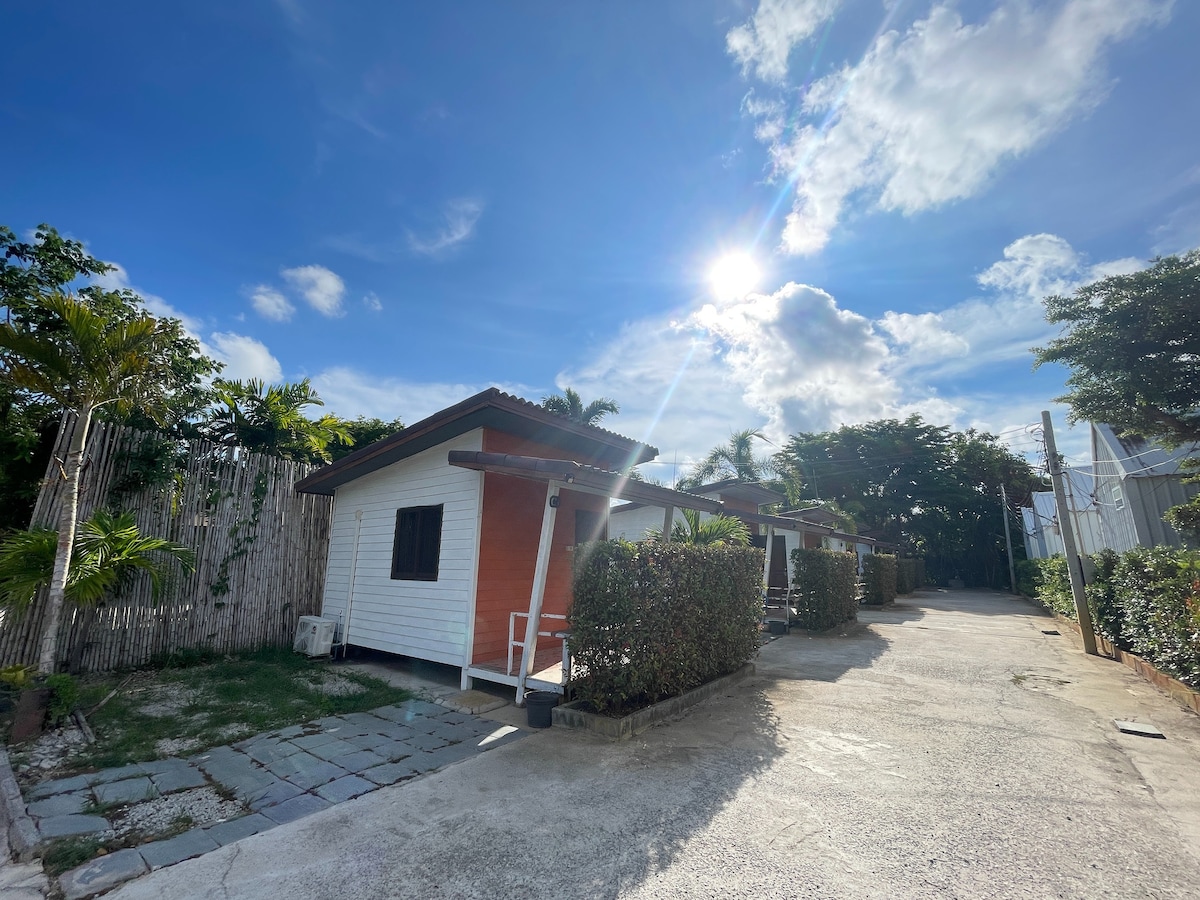 A row of charming single-story cabins features wooden accents and large windows, situated under a bright blue sky. Lush greenery and neatly trimmed hedges line the path, leading to the cabins. The sunlight creates a warm, inviting atmosphere throughout the space.