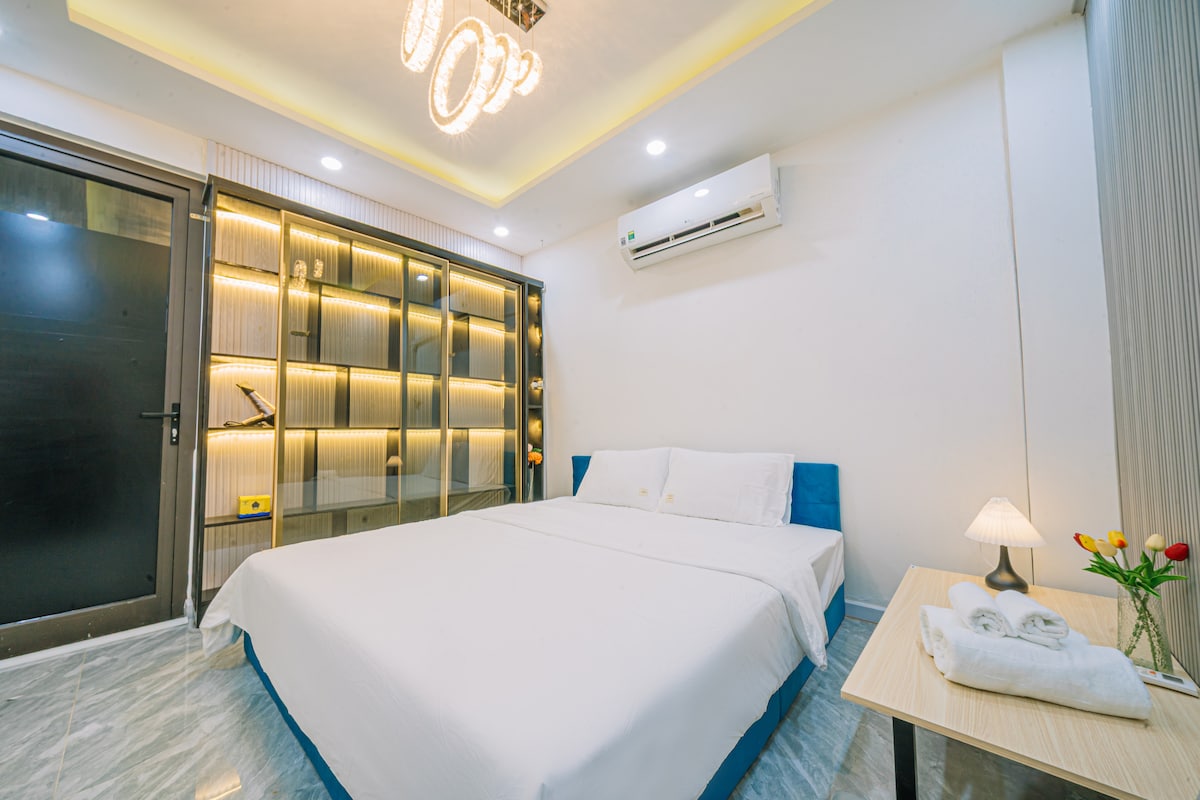 A spacious sleeping area is presented with a large bed covered in crisp white linens. Ambient lighting is provided by a modern chandelier and a bedside lamp. A glass cabinet with illuminated shelves displays decorative items, while a door leads to the exterior.