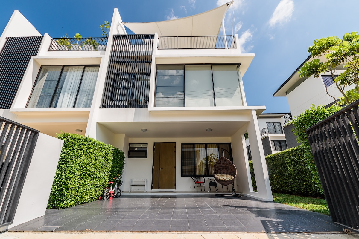 A modern townhouse exterior is presented, featuring clean lines and large windows that allow natural light. A shaded terrace is visible at the entrance, adorned with a hanging chair. Lush greenery surrounds the property, enhancing the inviting ambiance.