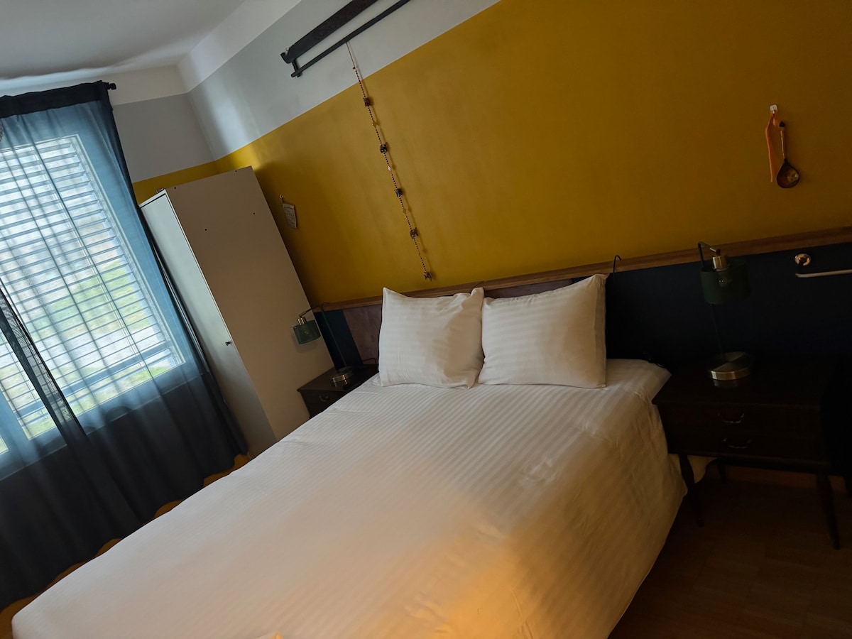 The room features a spacious bed with crisp white linens against a yellow and neutral wall. A window with sheer curtains allows natural light to filter in. Two bedside tables with lamps are positioned beside the bed, enhancing the serene atmosphere.