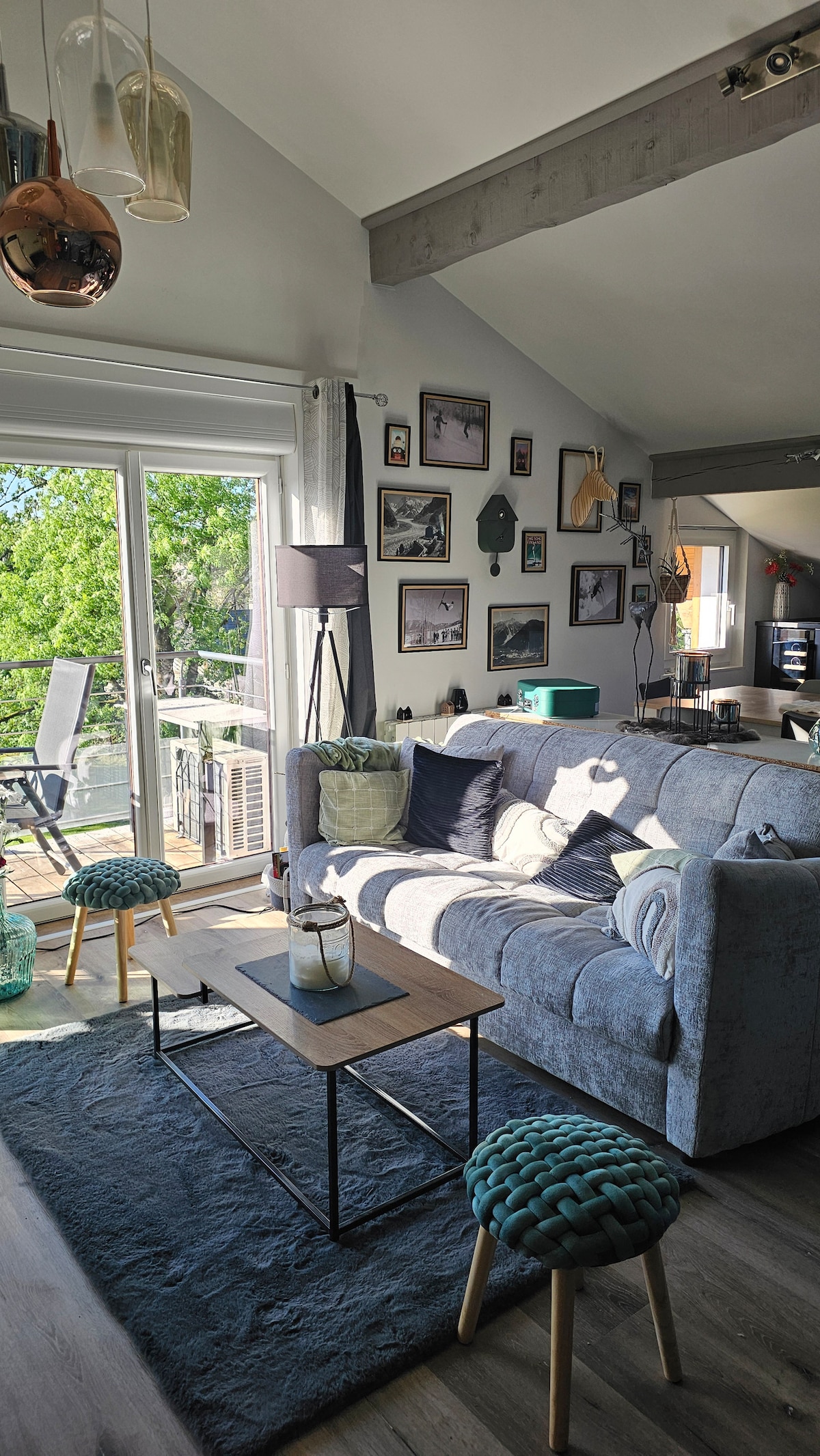 A comfortable living area features a sofa adorned with various cushions and a coffee table placed on a textured rug. Decorative wall art is displayed above, while sunlight fills the space through sliding glass doors that open to a small balcony overlooking greenery.