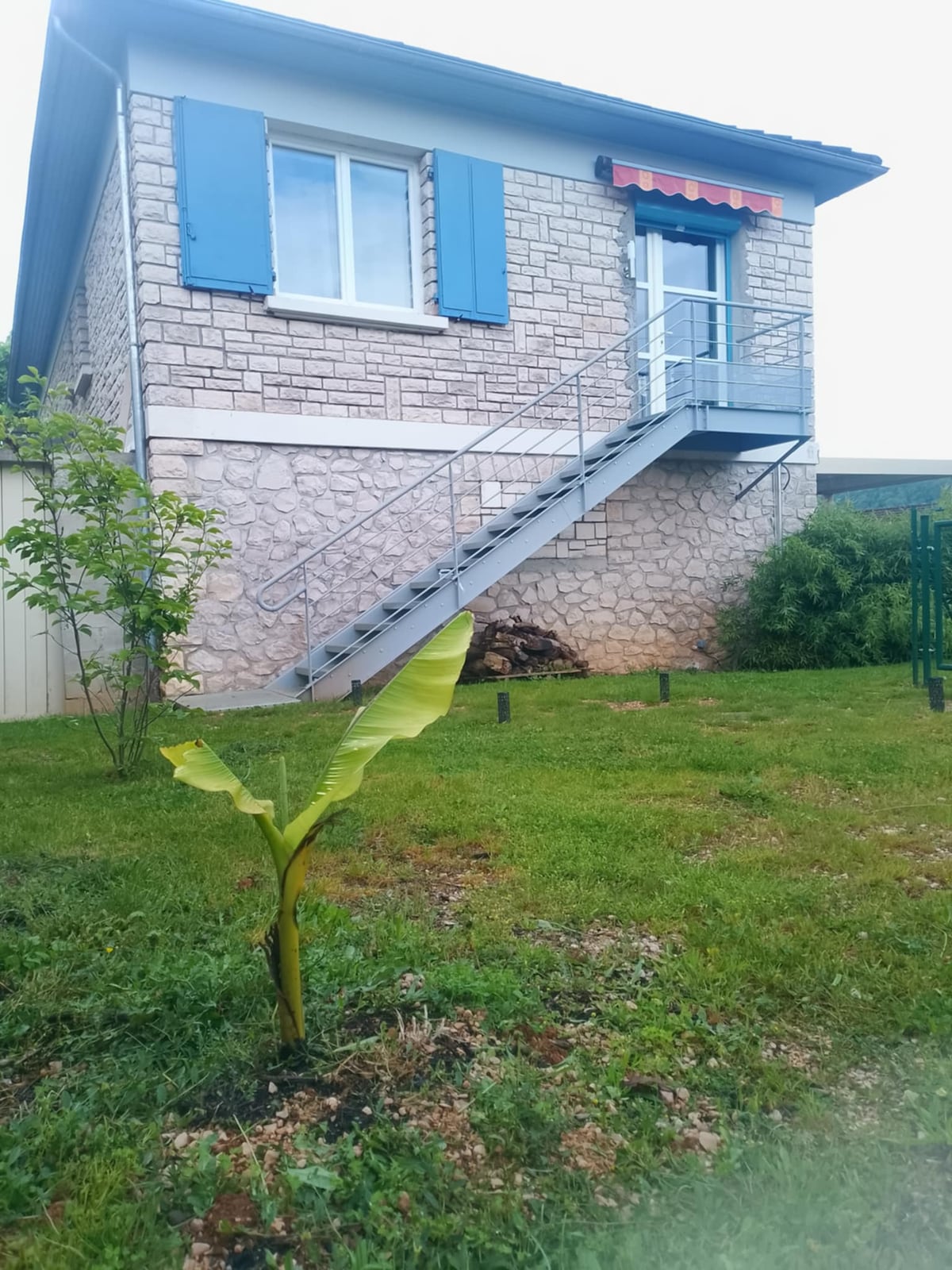 A two-story building is presented with a light stone facade and blue shutters. A metal staircase leads to an entrance adorned with an awning. A young banana plant is positioned in the grass in front of the building, which is surrounded by a landscaped area.
