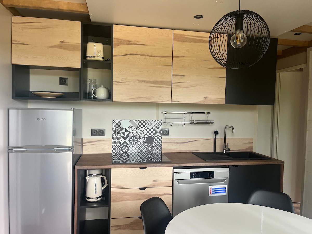 A modern kitchen area features an arrangement of light wood and dark cabinetry. Stainless steel appliances, including a refrigerator and dishwasher, are present. An electric kettle and other kitchen essentials are neatly organized on the countertop. A round dining table with seating is also visible.