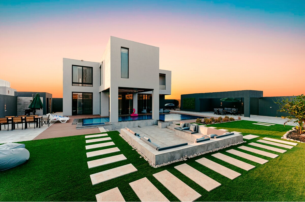 The villa's modern architecture is showcased against a vibrant sunset. An outdoor seating area is framed by the spacious lawn, with stone pathways leading to a private pool. Surrounding greenery and lounge areas provide a serene environment for relaxation.