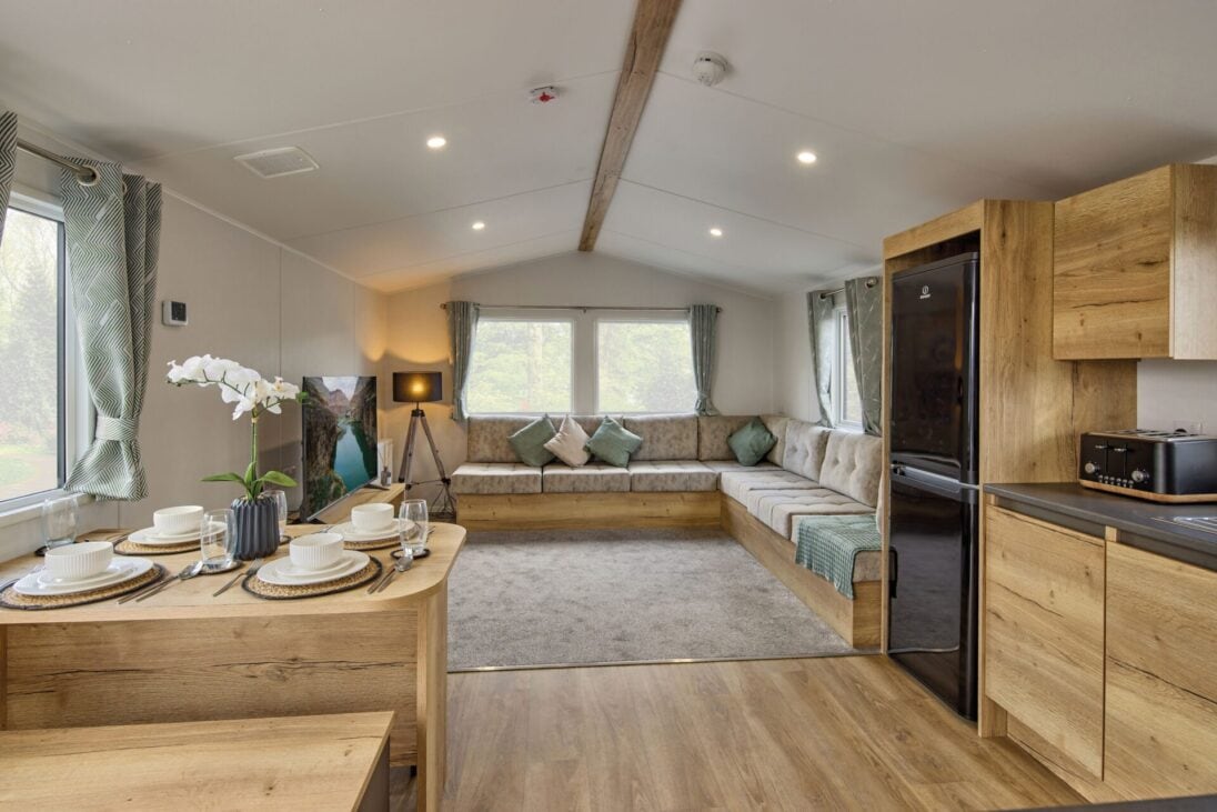 The open-plan living area features a spacious lounge with a comfortable sectional sofa, adorned with decorative cushions. A modern dining table is set with plates and utensils. Natural light filters through large windows, enhancing the wooden cabinetry and warm tones of the decor.