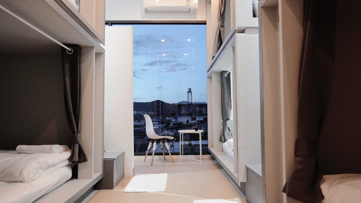 The interior of a shared dormitory is presented, showing multiple bunk beds arranged along the walls. A small table and a white chair are positioned near a large window, revealing a view of the outside landscape. Natural light is allowed in through the glass.