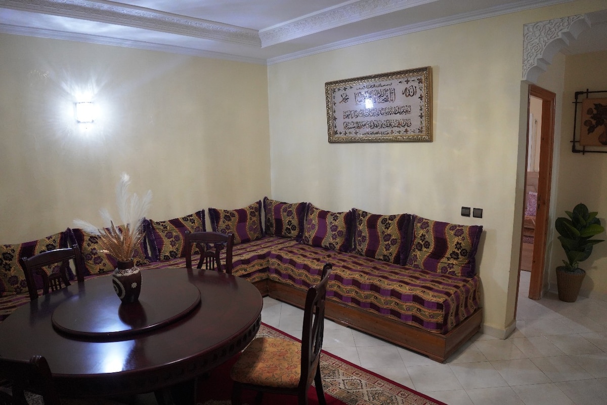 A spacious living area features a circular wooden table surrounded by a large cushioned seating area with intricate patterns. A decorative piece is placed at the center of the table, while a potted plant adds a touch of greenery. Soft lighting complements the warm wall tones.