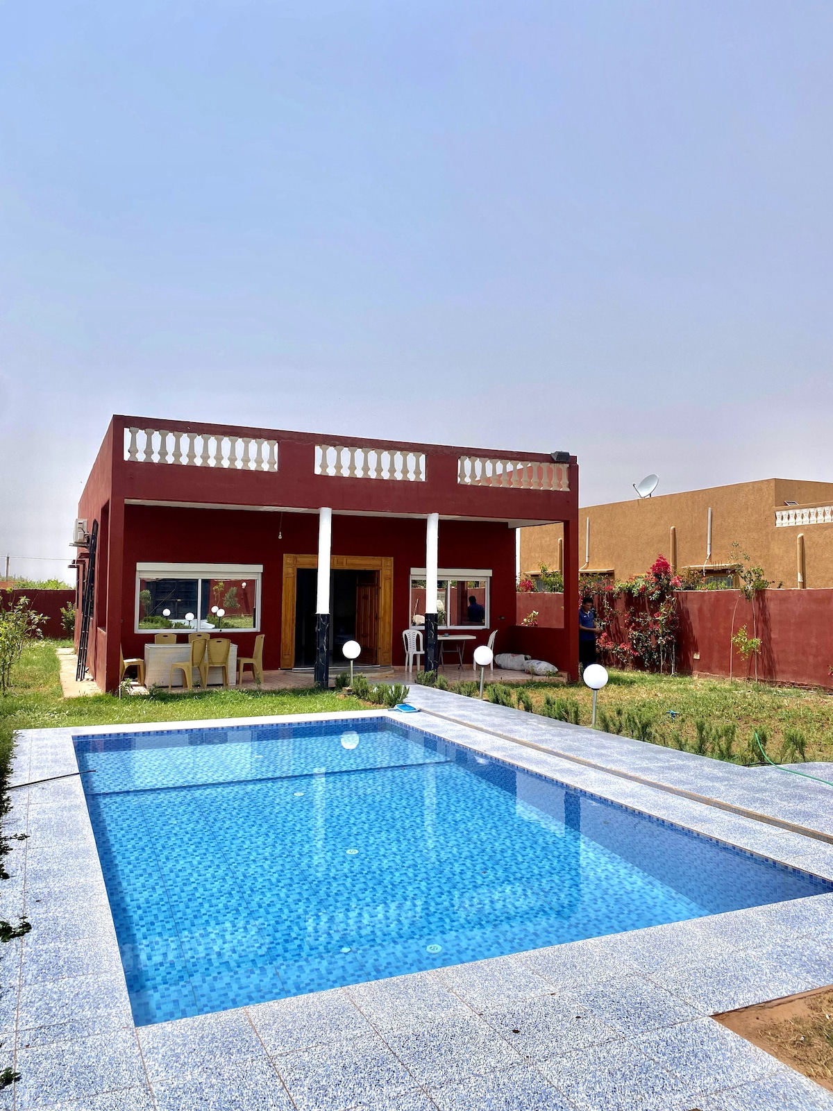 The image showcases a two-story villa with a spacious pool in the foreground. The pool, featuring a light blue tile border, is surrounded by a well-maintained garden. The villa is painted in a warm red hue, with large windows and a balcony adorned with white railings.