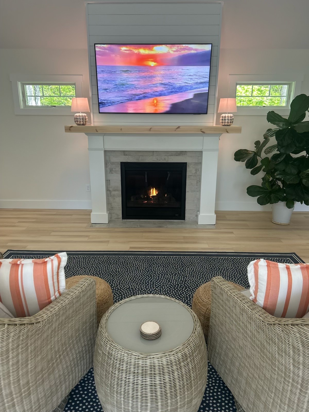 A welcoming living area is highlighted by a modern fireplace beneath a flat-screen TV displaying a sunset. Two comfortable chairs with striped cushions face a small table. Large windows allow natural light to fill the space, complemented by greenery from a nearby plant.