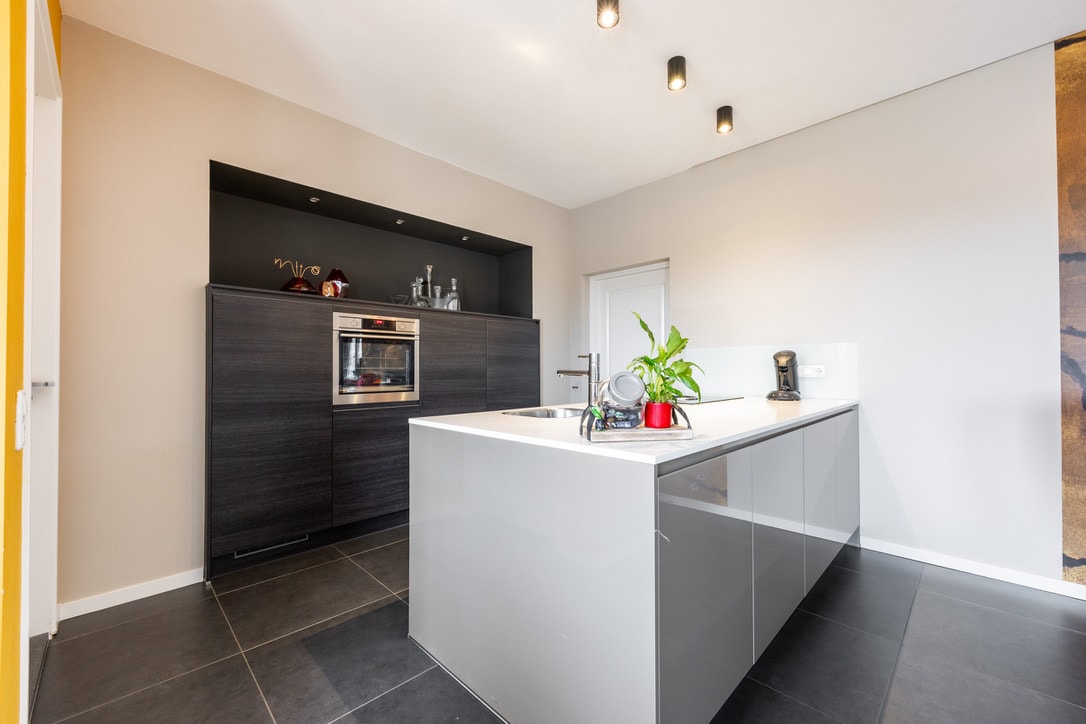A modern kitchen is showcased with a sleek island in the center, featuring a stovetop and sink. Dark cabinetry provides ample storage, complemented by built-in appliances, including an oven. A minimalist plant adds a touch of greenery, while the contemporary design emphasizes clean lines.