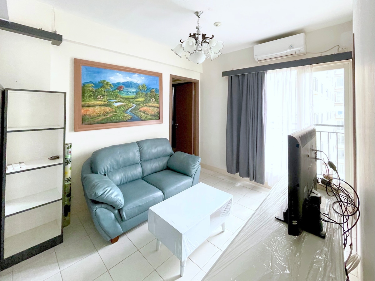 A living area is showcased with a light blue sofa facing a simple coffee table. A television set sits on a white console, while a painting of a landscape is displayed on the wall. Natural light enters through the balcony doors, enhanced by sheer gray curtains.