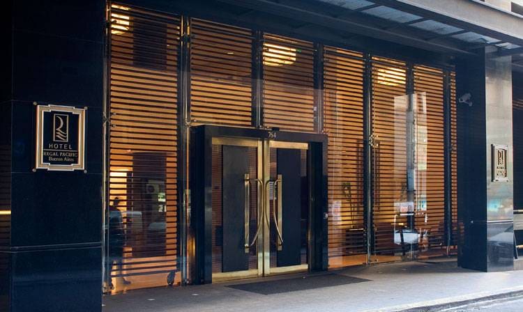The entrance to Hotel Regal Pacific is marked by glass doors framed in dark metal, surrounded by horizontal wooden slats. The sleek facade is complemented by warm lighting, creating a modern appeal. A discreet hotel logo is displayed prominently near the entrance.