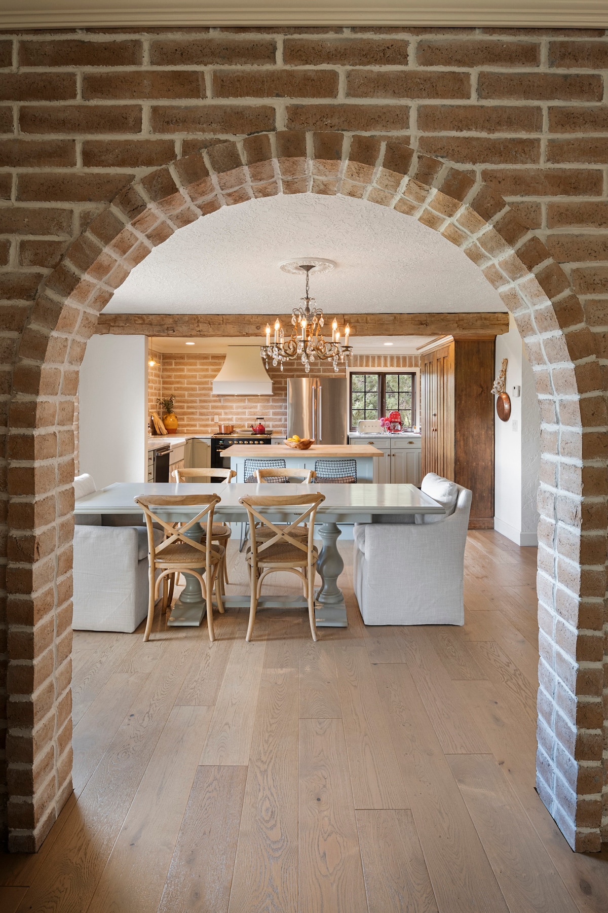An archway made of brick frames the view into the dining area, featuring a wooden table surrounded by six light-colored chairs. A chandelier hangs above the table, with the kitchen visible in the background showcasing modern appliances and natural light.
