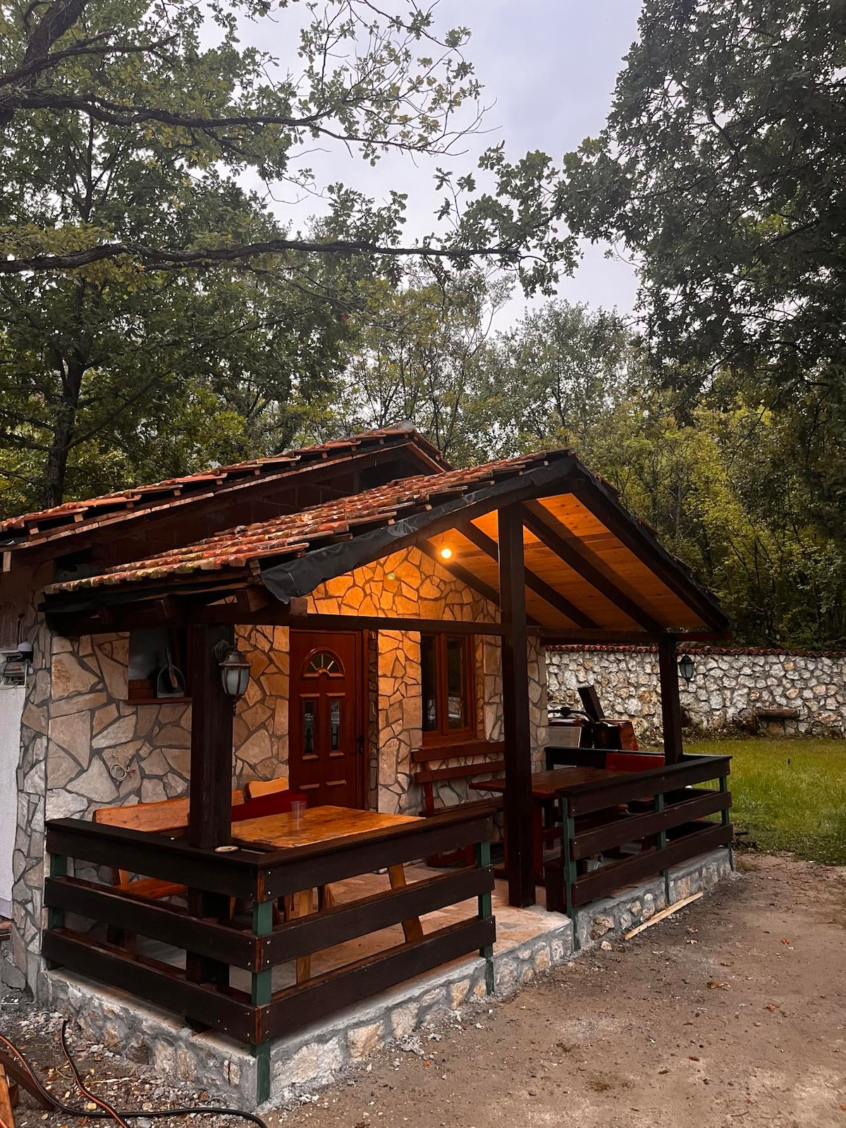 A quaint stone cottage features a wooden porch with an overhanging roof and warm lighting. The entrance is framed by a decorative door and windows, surrounded by lush greenery and a stone wall in the background. Natural elements enhance the peaceful ambiance.