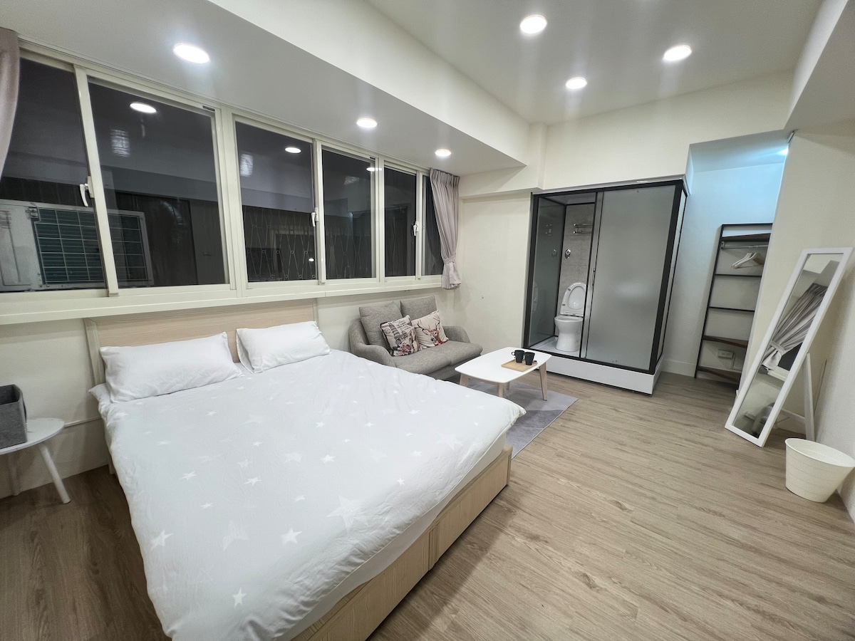 A bright and airy room features a spacious bed with a star-patterned coverlet. A cozy seating area is located beside large windows. A shower enclosure stands in the corner, and a mirror is positioned against the wall, enhancing the overall functionality of the space.